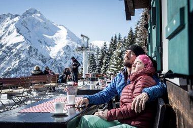 A couple relaxing with coffee at a mountain hut during winter holiday with snow-covered peaks in the background. | © Kleinwalsertal Tourismus eGen • Photographer: Dominik Berchtold