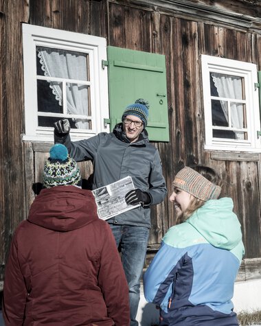 Fünf Personen vor einer alten Holzhütte im Winter, eine Person hält eine Zeitung, die anderen hören zu. | © Kleinwalsertal Tourismus eGen • Fotograf: Dominik Berchtold