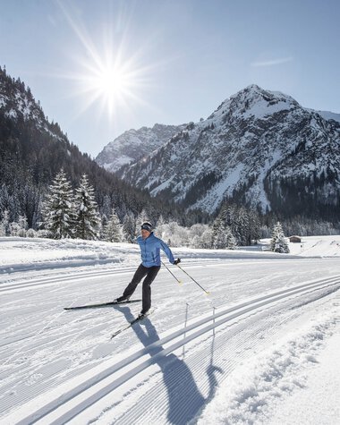Skifahrer auf Langlaufbahn in verschneiter Winterlandschaft mit Bergen und Sonne im Hintergrund. | © Kleinwalsertal Tourismus eGen • Fotograf: Dominik Berchtold