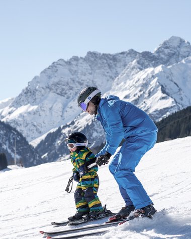 Ein Erwachsener hilft einem Kind beim Skifahren auf schneebedelter Piste vor einer beeindruckenden Bergkulisse. | © Kleinwalsertal Tourismus eGen • Fotograf: Dominik Berchtold