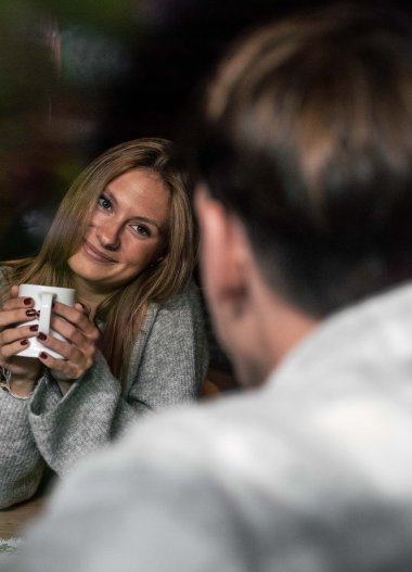 A young woman holding a mug and smiling warmly while sitting across from a man, in a cozy, private setting. | © Kleinwalsertal Tourismus eGen • Photographer: Hannah Bichay