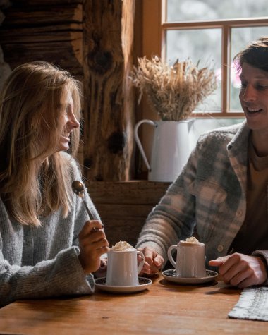 Two young people sit across from each other in a cozy café, laughing and enjoying cups of coffee in a rustic wooden and stone setting illuminated by candlelight. | © Kleinwalsertal Tourismus eGen • Photographer: Hannah Bichay