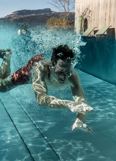 A man is diving into a swimming pool with splashing water, mountains, and a pavilion in the background. | ©  Kleinwalsertal Tourismus eGen • Photographer: Hannah Bichay