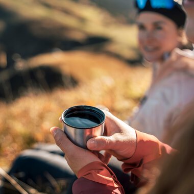 Zwei Menschen beim Wandern in der Natur, eine Person hält eine Kaffeetasse, im Hintergrund sanfte Landschaft bei Tageslicht. | © Kleinwalsertal Tourismus eGen • Fotograf: Hannah Bichay