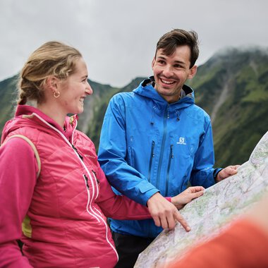 Two hikers with backpacks, laughing and chatting during an autumn hike in Kleinwalsertal, with mountain scenery in the background. | © Kleinwalsertal Tourismus eGen • Photographer: Oliver Farys