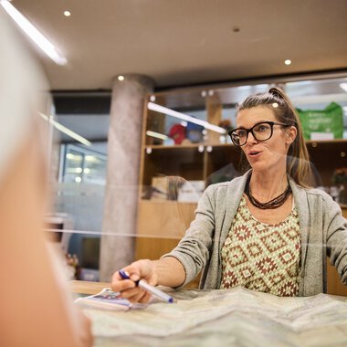 Frau mit Brille und lockigem Haar sitzt an einem Tisch in einem Kundenservice-Büro, spricht freundlich und hat Papiere vor sich. | © Kleinwalsertal Tourismus eGen • Fotograf: Oliver Farys