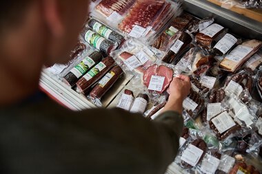 Eine Person wählt an einem Metzergeschäftsstand im Kleinwalsertal aus einer Vielzahl von vakuumverpacktem Fleisch und Wurstwaren. | © Kleinwalsertal Tourismus eGen • Fotograf: Oliver Farys