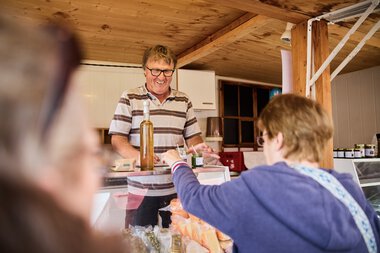 A man with glasses and a striped shirt smiling at the Walser Weekly Market in Kleinwalsertal summer 2023, as a woman hands him a product. | © Kleinwalsertal Tourismus eGen • Photographer: Oliver Farys