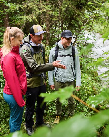 Three people, including a ranger, conducting an outdoor educational session in a green forest by a river. | © Kleinwalsertal Tourismus eGen • Photographer: Oliver Farys