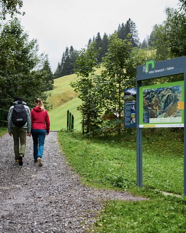 Zwei Personen wandern auf einem Waldweg im Kleinwalsertal, vorbei an Informationsschildern und umgeben von Bäumen. | © Kleinwalsertal Tourismus eGen • Fotograf: Oliver Farys