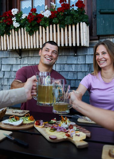 Four friends cheers with beer mugs during an outdoor meal, with a variety of snacks on the table, in front of a traditional alpine building decorated with flower boxes. | © Kleinwalsertal Tourismus eGen • Photographer: Oliver Farys