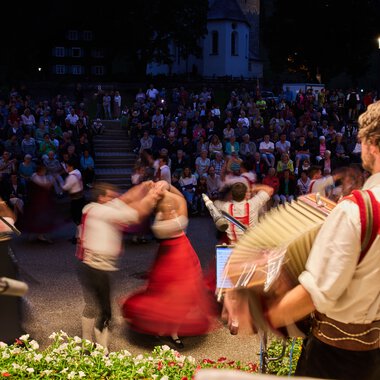 Musiker in traditioneller Tracht spielt bei einem Sommer-Festival im Kleinwalsertal, während im Hintergrund eine große Menschenmenge tanzt und zuschaut. | © Kleinwalsertal Tourismus eGen • Fotograf: Oliver Farys