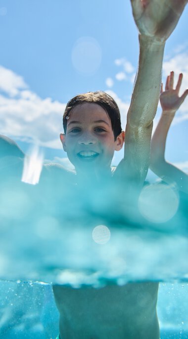 Two children swimming and smiling in the outdoor pool at Freibad Riezlern, with a bright blue sky and clouds in the background. | © Kleinwalsertal Tourismus eGen • Photographer: Oliver Farys