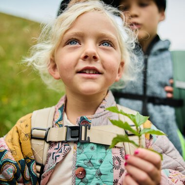 Kinder beim Naturerlebnis im Sommer 2024, ein Mädchen mit Rucksack hält eine Pflanze, umgeben von grüner Landschaft. | © Kleinwalsertal Tourismus eGen • Fotograf: Oliver Farys