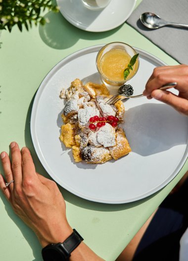 Table with a plate of delicious Kaiserschmarrn dusted with powdered sugar and garnished with cherries, alongside a glass of fresh orange juice. | © Kleinwalsertal Tourismus eGen • Photographer: Oliver Farys