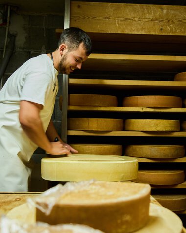 Man in a white coat working in a cheese cellar or cheese-making environment, surrounded by round cheeses aging on shelves. | © Kleinwalsertal Tourismus eGen • Photographer: Oliver Farys