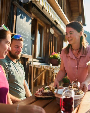 Three people enjoying summer outdoors at a mountain hut in Kleinwalsertal, sharing meals and drinks on a wooden tray. | © Kleinwalsertal Tourismus eGen • Photographer: Oliver Farys