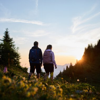 Ein Paar wandert bei Sonnenaufgang durch die Natur im Kleinwalsertal, umgeben von Bäumen und Blumen, mit Blick auf den Himmel und die aufgehende Sonne. | © Kleinwalsertal Tourismus eGen • Fotograf: Oliver Farys