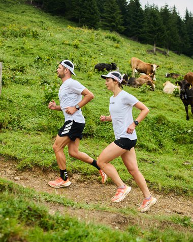 Two women running on a grassy trail surrounded by cows and a forest in the background. | © Kleinwalsertal Tourismus eGen • Photographer: Oliver Farys