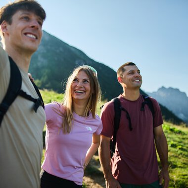 Three young adults hiking in the mountains, smiling and enjoying nature in Kleinwalsertal. | © Kleinwalsertal Tourismus eGen • Photographer: Oliver Farys