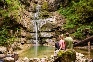 Two people sitting by the waterfall in Kleinwalsertal surrounded by green nature, overlooking the waterfall and the calm water. | © Kleinwalsertal Tourismus eGen • Photographer: Oliver Farys