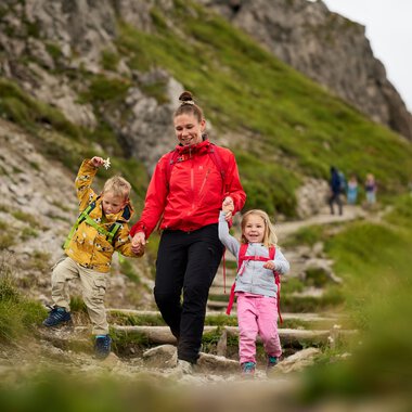 Familie beim Wandern in den Bergen, Kinder und Erwachsene auf einem Pfad mit grüner Berghänge im Hintergrund. | © Kleinwalsertal Tourismus eGen • Fotograf: Oliver Farys
