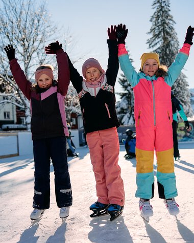 Kinder beim Eislaufen auf einem zugefrorenen See im Winter, fröhlich mit erhobenen Armen, umgeben von verschneiten Bäumen und anderen Eisläufern. | © Kleinwalsertal Tourismus eGen • Fotograf: Oliver Farys