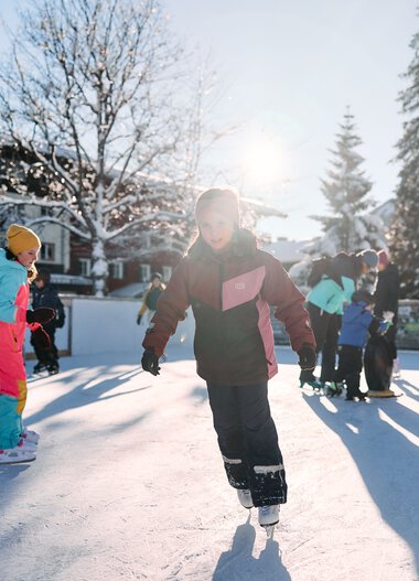 Children ice skating on an outdoor ice rink in winter, surrounded by trees and buildings, sunny day. | © Kleinwalsertal Tourismus eGen • Photographer: Oliver Farys