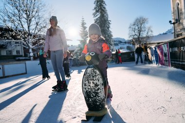Kinder beim Eislaufen auf dem Eisplatz in Riezlern im Winter, umgeben von Schneelandschaft und wenigen Erwachsenen. | © Kleinwalsertal Tourismus eGen • Fotograf: Oliver Farys