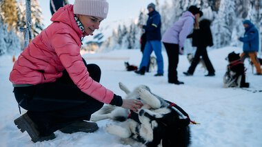 Young woman playing with a husky sled dog in snowy winter landscape, with people in winter activity gear in the background. | © Kleinwalsertal Tourismus eGen • Photographer: Oliver Farys