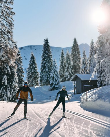 Zwei Skifahrer ziehen ihre Spuren im verschneiten Winterwald beim Langlaufen im Kleinwalsertal, Sonnenschein und klare Sicht. | ©  Kleinwalsertal Tourismus eGen • Fotograf: Oliver Farys
