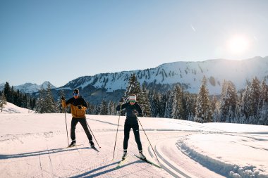 Two cross-country skiers in a winter landscape in Kleinwalsertal, surrounded by snow-covered trees and mountains, sunny weather. | © Kleinwalsertal Tourismus eGen • Photographer: Oliver Farys