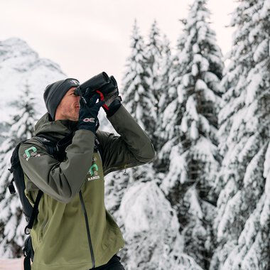 Mann mit Winterbekleidung und Fernglas in verschneitem Waldgebiet in den Alpen, wahrscheinlich ein Ranger oder Naturführer. | © Kleinwalsertal Tourismus eGen • Fotograf: Oliver Farys