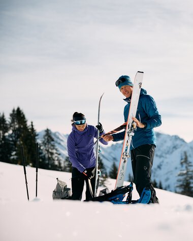 Two skiers on a snowy mountain landscape, one helping the other with ski equipment, with mountains and pine trees in the background. | © Kleinwalsertal Tourismus eGen • Photographer: Oliver Farys