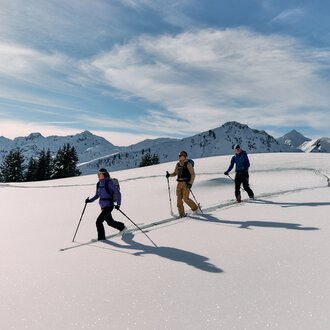Three ski tourers in snow-covered mountain landscape with peaks and cloudy sky during winter tour in Kleinwalsertal. | © Kleinwalsertal Tourismus eGen • Photographer: Oliver Farys