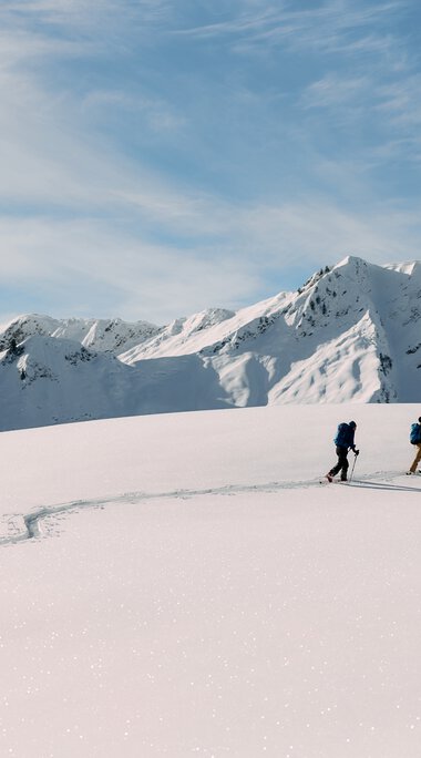 Three ski tourers traveling across snow-covered landscape in the Kleinwalsertal, surrounded by high, snow-capped peaks on a clear day. | © Kleinwalsertal Tourismus eGen • Photographer: Oliver Farys