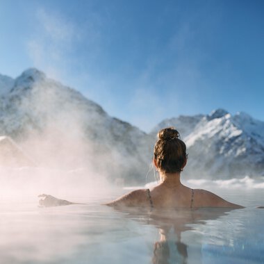 Woman relaxing in a warm spa pool with snowy mountains in the background in Kleinwalsertal, surrounded by mist and nature. | © Kleinwalsertal Tourismus eGen • Photographer: Oliver Farys