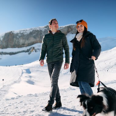 Two people hiking in snowy weather in Kleinwalsertal, accompanied by a dog, with mountains in the background. | © Kleinwalsertal Tourismus eGen • Photographer: Oliver Farys