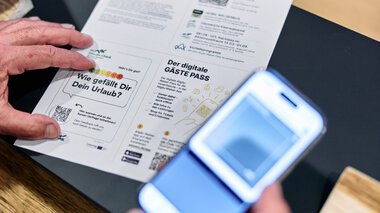 Person holds a digital guest pass and reads a printed document at a table, while someone takes a photo of it with a smartphone. | © Kleinwalsertal Tourismus eGen • Photographer: Oliver Farys