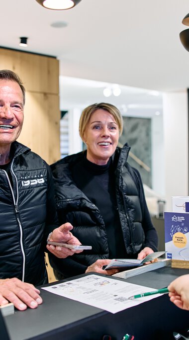 Two smiling people at the reception desk during winter 2025 registration at Kleinwalsertal, served by an employee. | © Kleinwalsertal Tourismus eGen • Photographer: Oliver Farys