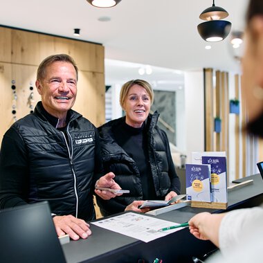 Zwei lachende Personen am Empfang bei einer Registrierung im Winter 2025 im Kleinwalsertal, betreut von einem Mitarbeiter. | © Kleinwalsertal Tourismus eGen • Fotograf: Oliver Farys