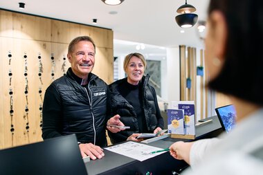 Two smiling people at the reception desk during winter 2025 registration at Kleinwalsertal, served by an employee. | © Kleinwalsertal Tourismus eGen • Photographer: Oliver Farys