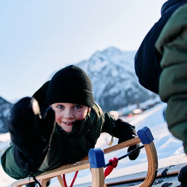Child sledding in Kleinwalsertal, smiling and wearing winter clothes, with mountains in the background. | © Kleinwalsertal Tourismus eGen • Photographer: Oliver Farys