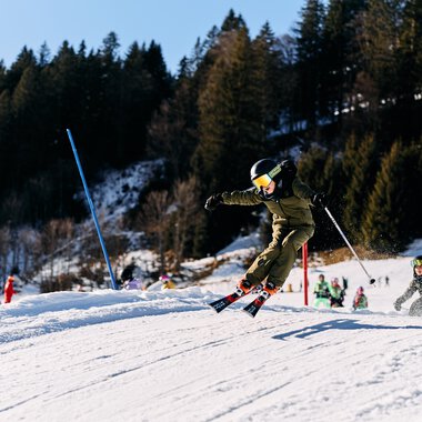 Kinder beim Skifahren auf einer Skipiste im Kleinwalsertal, umgeben von Bäumen und winterlicher Landschaft, bei Sonnenschein. | © Kleinwalsertal Tourismus eGen • Fotograf: Oliver Farys