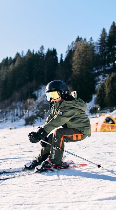 A child skiing in Kleinwalsertal in winter 2025, wearing a helmet and ski gear, with snow-covered landscape and trees in the background. | © Kleinwalsertal Tourismus eGen • Photographer: Oliver Farys
