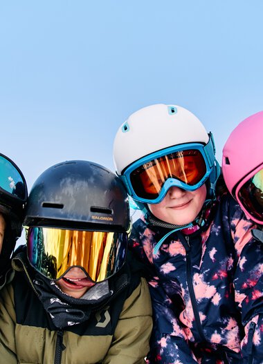 Vier Kinder beim Skifahren in Winterkleidung und Helmen, lachen und posieren im Schnee. | © Kleinwalsertal Tourismus eGen • Fotograf: Oliver Farys