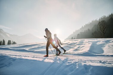 Two cross-country skiers on the Schwarzwasserloipe in Kleinwalsertal during sunrise, surrounded by snowy landscape and mountains. | © Kleinwalsertal Tourismus eGen • Photographer: Oliver Farys