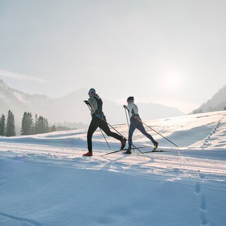 Two cross-country skiers skiing on the snowy Schwarzwasserloipe trail in Kleinwalsertal during sunrise, surrounded by mountains and pine trees. | © Kleinwalsertal Tourismus eGen • Photographer: Oliver Farys