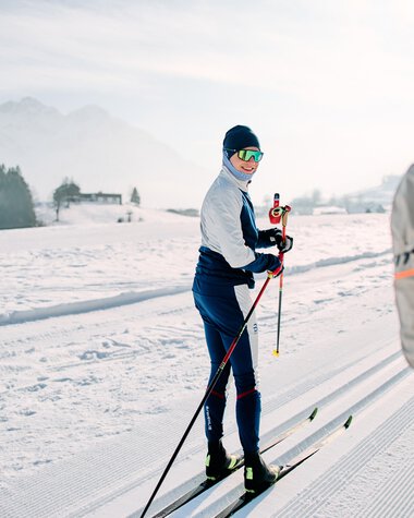 Cross-country skier on a snowy trail with mountains and winter landscape in the background, talking to someone in the foreground holding ski poles. | © Kleinwalsertal Tourismus eGen • Photographer: Oliver Farys