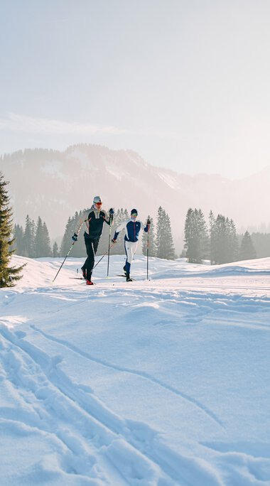 Two people cross-country skiing in a snowy landscape with a wooden cabin and pine trees in the background. | © Kleinwalsertal Tourismus eGen • Photographer: Oliver Farys